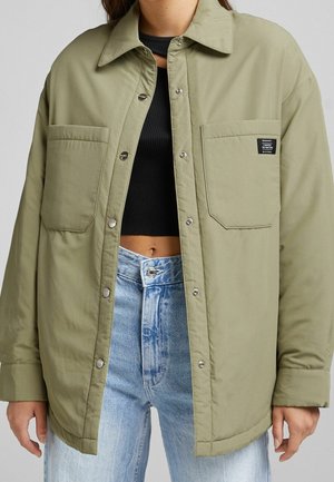 Kvinde iført en oversized olivengrøn jakke med to store brystlommer over en sort crop top og lyseblå højtaljede jeans.