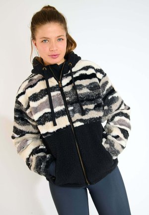 Giacca in pile con un motivo a righe in nero, bianco e grigio. Ha un cappuccio, chiusura con zip e tasche laterali. Texture morbida.