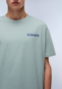 Tříčtvrteční světle tyrkysové tričko z bavlny. Na levé části hrudi má modré logo a kulatý výstřih. Hladká textura.