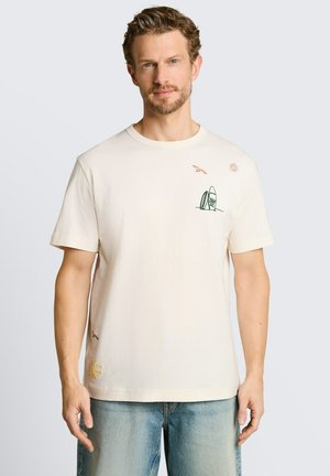 Mężczyzna z lekkim zarostem, noszący kremową koszulkę z małymi wzorami deski surfingowej, ptaka i słońca, zestawioną z jasnoniebieskimi dżinsami.