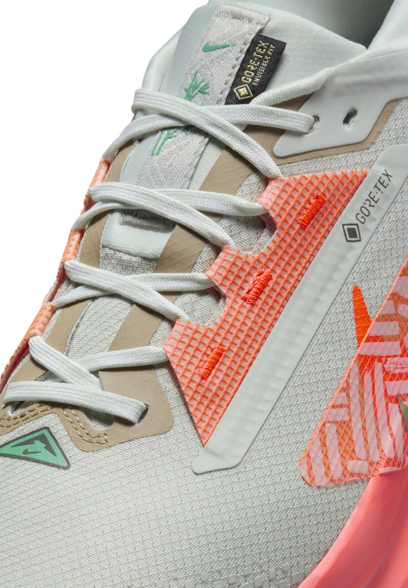 Zapatilla gris claro con una superficie texturizada, que presenta detalles en naranja y una etiqueta de Gore-Tex. Incluye cordones blancos y un diseño aerodinámico.