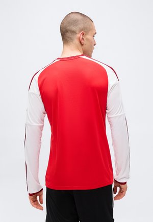 Homme aux cheveux courts portant un maillot de sport rouge et blanc à manches longues et un pantalon noir, de dos sur un fond gris uni.