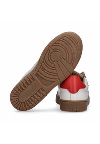 Sneaker bianco con accento rosso sul tallone, suola in gomma marrone con battistrada decorato e borchie metalliche lungo il lato.