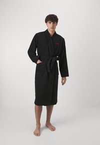 Peignoir de bain en tissu éponge noir avec un col châle, des manches longues, une ceinture à la taille et deux poches avant ; arbore le logo rouge "HUGO" sur la poitrine.