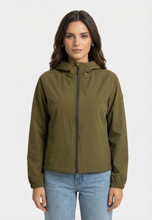 Donna con giacca con cappuccio verde oliva con cerniera frontale e jeans azzurri, in piedi contro uno sfondo bianco uniforme.