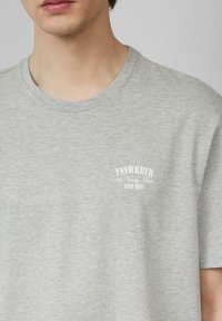Lichtgrijze katoenen t-shirt met korte mouwen, ronde halslijn en witte geprinte tekst op de linkerborst. Zachte stof, normale pasvorm.