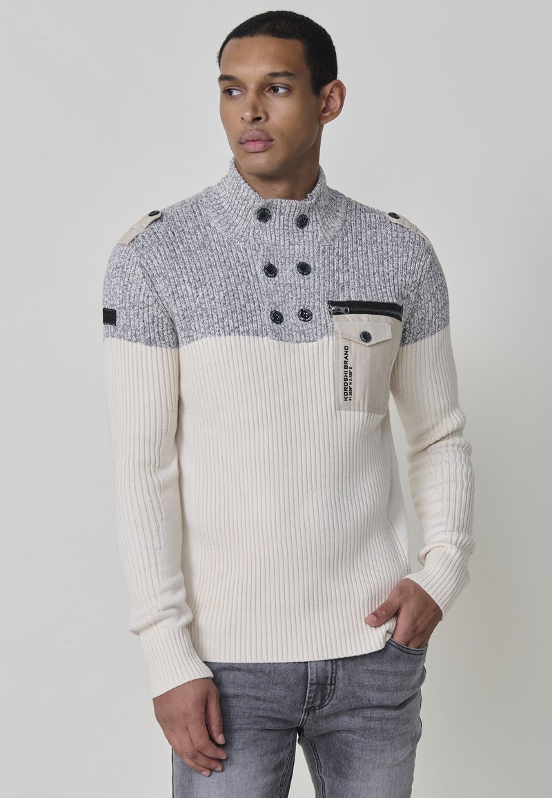 Suéter de punto con textura acanalada, bloques de color gris y crema, cuello de embudo, cuatro botones negros y un bolsillo con cremallera con un acento de etiqueta.