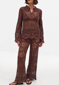 Brązowy, szydełkowany zestaw składający się z dwóch części, składający się z koszuli zapinanej na guziki w kwiatowe wzory oraz spodni o szerokich nogawkach. Przezroczysta struktura z misternym wzorem koronkowym.