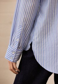 Mėlyna ir balta dryžuota marškinėliai su sagotu rankogaliu, pagaminti iš lengvos medžiagos, su išlenktu kraštu ir lygiu paviršiumi.