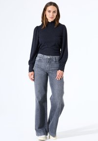 Dunkelblaue gerippte Strickoberteil mit hohem Kragen und gepufften Ärmeln, kombiniert mit hellgrauen Schlagjeans und cremefarbenen Schuhen.