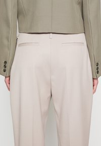 Ušitý blejzr v olivové zelené barvě v kombinaci s béžovými kalhotami s vysokým pasem. Kalhoty mají hladký povrch a dvě zadní kapsy.