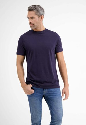 Mann mit grauem Haar, der ein dunkelblaues kurzärmliges T-Shirt und blaue Jeans trägt, zur Seite blickt und eine Hand in seiner Tasche hat.