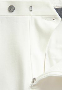 Weiße Baumwollhosen verfügen über einen Knopfverschluss, einen Reißverschluss und eine glatte Textur. Die Nähte sind an den Abschlüssen sichtbar, um zusätzliche Haltbarkeit zu gewährleisten.