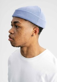 Lichtblauwe gebreide beanie met een geribbelde textuur, schuin op het hoofd gedragen. Witte crewneck t-shirt. Close-up profielweergave.