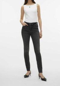 Jeans skinny de denim negro con una cintura alta, combinados con una blusa sin mangas blanca y tacones negros de punta afilada, con un diseño elegante.