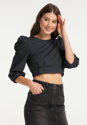 Jonge vrouw lacht, draagt een zwart cropped top met pofmouwen en een high-waisted zwarte denim rok met ritsdetail, hand in de zak.