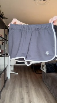Harmaat urheilushortsit, joissa on valkoinen reunus, elastinen vyötärönauha ja pieni logo. Kangas näyttää pehmeältä ja kevyeltä, suunniteltu mukavuutta varten.