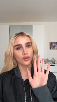 Une jeune femme aux longs cheveux blonds et portant une veste noire lève sa main gauche avec les doigts étendus dans une pièce bien éclairée.