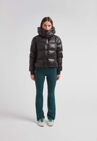 Chaqueta de plumas negra con un cuello alto, textura brillante y cremallera frontal. Llevada sobre pantalones ajustados verdes y zapatos blancos con detalles verdes.