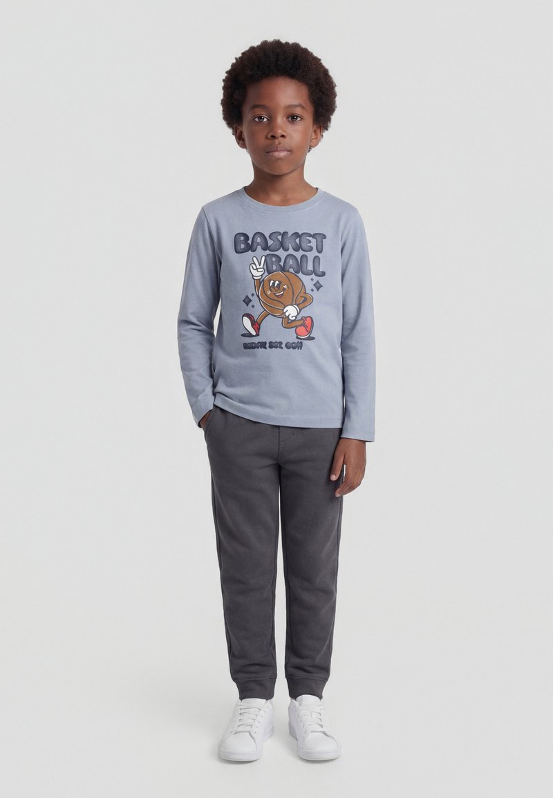 Garçon debout portant une chemise à manches longues bleu clair avec un dessin animé de basketball et un pantalon foncé, baskets blanches sur un fond uni.