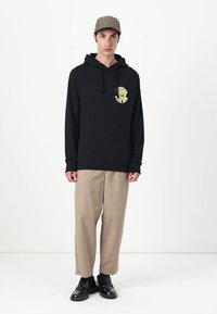 Felpa nera con cappuccio e una grafica colorata sul petto, abbinata a pantaloni beige e scarpe nere, con un taglio comodo e tasche.