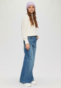 Lichte crème trui, losse pasvorm, ribbeltextuur. Blauwe denim wijde pijpen jeans met verticale witte strepen. Witte sneakers en lavendel beanie.