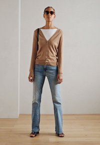Gilet marron clair avec des boutons, haut blanc en dessous, jean bleu clair et lunettes de soleil noires. Debout sur un sol en bois devant un mur uni.