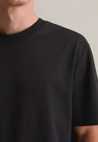 Muž nosí jednoduché čierne tričko s okrúhlym výstrihom, je vidieť krk, plece a hornú časť ramena na neutrálnej pozadí.