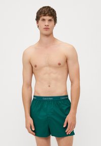 Zelene kopalne hlače iz gladkega materiala, z elastičnim pasom in logotipom "Calvin Klein". Klasičen kroj, dolžina do kolen.
