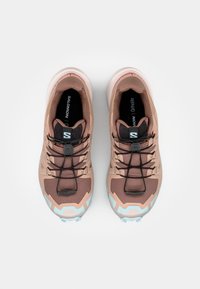 Pantofi sport într-o nuanță de maro deschis, cu un superior texturat, șireturi negre și o talpă amortizată cu accente de albastru deschis și un logo.