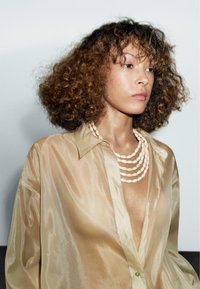 Transparante beige blouse met een diepe V-hals, gecombineerd met een meerlagige beige kralenketting. Krullend haar accentueert de zachte textuur.