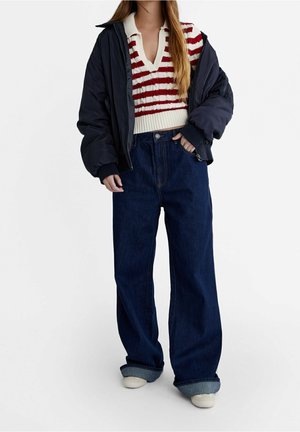 Femme portant un jean bleu marine ample, retroussé aux poignets, un top en maille rayé rouge et blanc, et une veste noire oversize sur fond uni.