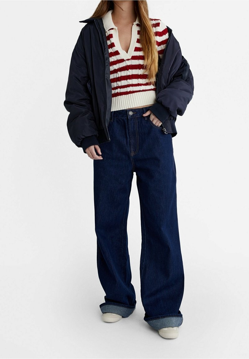 Femme portant un jean bleu marine ample, retroussé aux poignets, un top en maille rayé rouge et blanc, et une veste noire oversize sur fond uni.