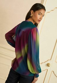 Wielokolorowa bluzka wykonana z lekkiego materiału, z długimi bufkowymi rękawami, falbaniastym dekoltem oraz gradientowym wzorem w odcieniach turkusu, fioletu i zieleni.