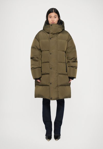 Cappotto in nylon verde oliva imbottito con cappuccio, grandi sezioni trapuntate, bottoni frontali e tasche laterali. Indossato su jeans scuri e scarpe a punta.