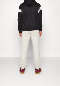 Black hooded jacket with white accents, light beige pants, and red sneakers with a gum sole, shot from the back against a white background.