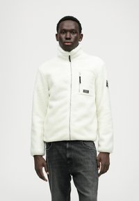 Veste en polaire blanche avec une fermeture éclair à l'avant, une poche poitrine zippée et des accents noirs. Associée à un jean gris foncé. Texture douce et design confortable.