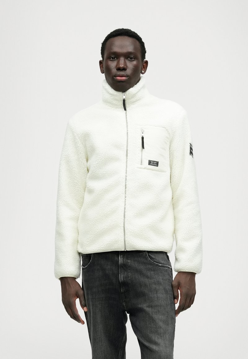 Veste en polaire blanche avec une fermeture éclair à l'avant, une poche poitrine zippée et des accents noirs. Associée à un jean gris foncé. Texture douce et design confortable.