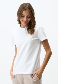 T-shirt bianco in cotone con maniche corte, scollatura rotonda, vestibilità comoda, texture liscia, indossato con pantaloni grigi chiari dotati di tasche laterali.