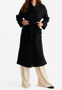 Dlouhý černý kabát z vlny, s širokými rukávy, páskem v pase a designem límce. V kombinaci s krémovými kalhotami.
