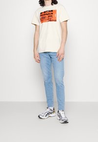 Camiseta beige con estampado gráfico en negro y naranja, combinada con jeans de colores azul claro y zapatos deportivos grises con detalles en negro.