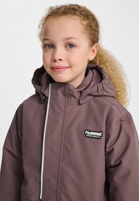 Lila wasserdichte Jacke mit Kapuze, Frontreißverschluss und weißen Akzenten. Verfügt über ein schwarzes Logo auf der Brust für verbesserte Sichtbarkeit.