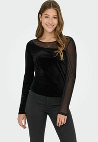 Mladá žena s dlouhými blond vlasy, oblečená v černém sametovém topu s průhlednými dlouhými rukávy a černými kalhotami, se usmívá na jednotném pozadí.