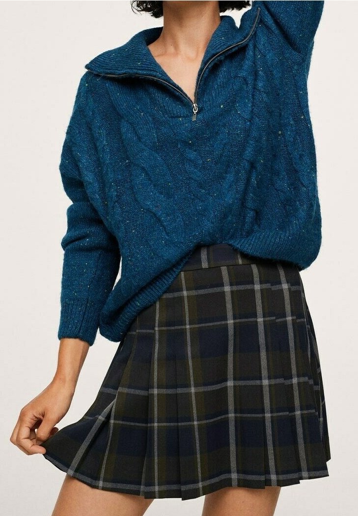 Personne portant un pull bleu en tricot torsadé avec une fermeture éclair partielle et une jupe plissée à carreaux foncés, tenant un côté de la jupe.