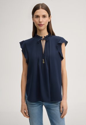 Kobieta o prostych brązowych włosach, ubrana w granatową bluzkę bez rękawów z falbankami na ramionach i wiązaniem przy szyi, w połączeniu z jasnoniebieskimi dżinsami.