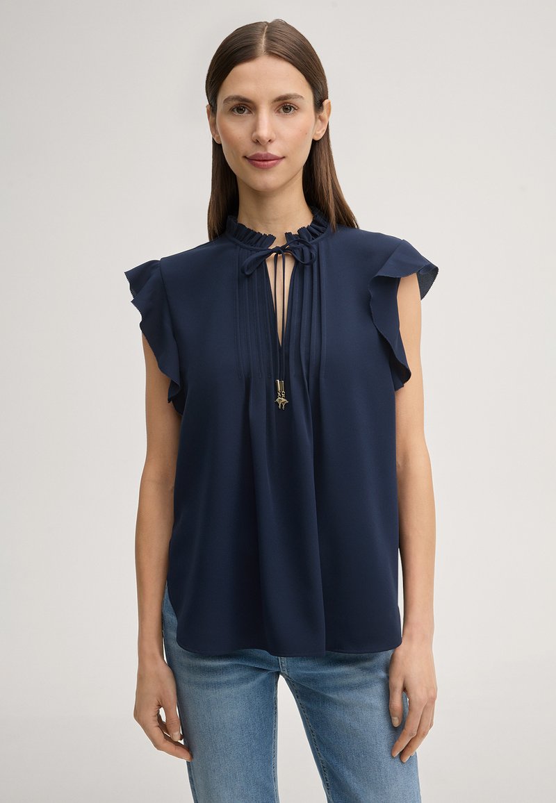 Frau mit glatten braunen Haaren, die eine ärmellose marineblaue Bluse mit Rüschenschultern und einer Schleifendetail am Kragen trägt, kombiniert mit hellblauen Jeans.