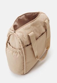 Beige mjuk tygväska med breda handtag, öppen toppdragkedja och samlade sidofickor, visad mot en enkel vit bakgrund.