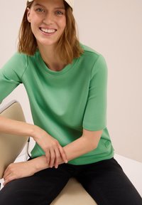 Femme souriante avec des cheveux jusqu'aux épaules portant une chemise verte à manches courtes, un pantalon noir et une casquette de couleur claire, assise sur une chaise beige.
