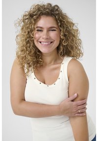 Débardeur couleur crème avec une texture côtelée, agrémenté de clous argentés autour de l'encolure. Le mannequin a les cheveux bouclés et sourit.