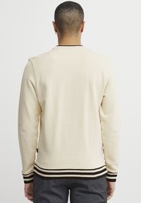 Sudadera de manga larga color crema con puños y dobladillo de canalé, que presenta rayas marrón oscuro; textura suave y diseño clásico de cuello redondo.